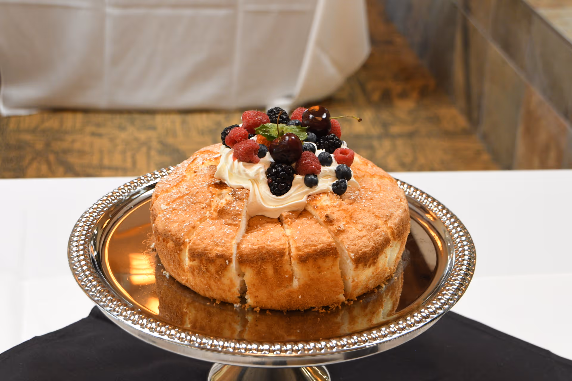 A round cake with a light brown crust topped with whipped cream and an assortment of fresh berries including raspberries, blackberries, blueberries, and cherries, placed on a silver cake stand with a decorative edge on a white tablecloth.