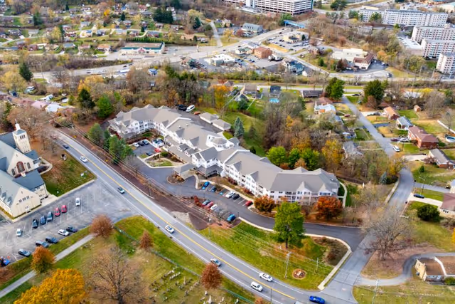 Aerial view of the Bethel Park senior living complex with connected buildings, parking lots, nearby church, roads, and surrounding neighborhood.