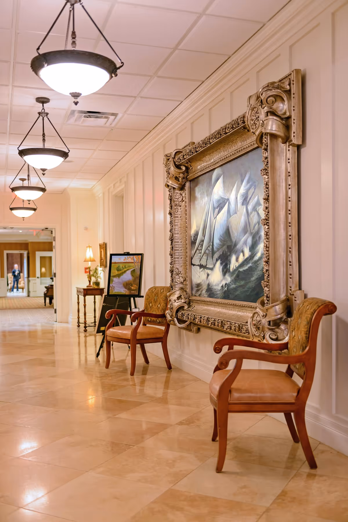 A well-lit hallway with beige tiled floors and white paneled walls. Two wooden chairs with cushioned seats are placed against the wall, flanking a large ornate framed painting of ships at sea. Further down the hallway, there is a small table with a lamp and a framed painting on an easel. Ceiling lights hang in a row along the corridor.
