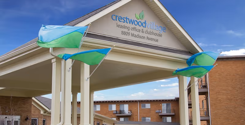 Entrance canopy of Crestwood Village leasing office and clubhouse with three blue and green flags flying, brick buildings in the background under a partly cloudy sky.