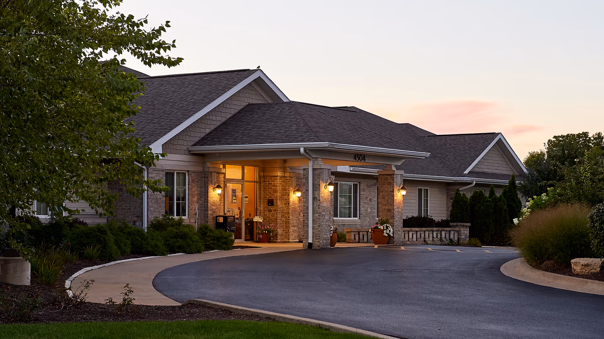 Exterior view of a single-story senior living facility building at dusk with warm lights illuminating the entrance. The building has a covered driveway, brick and siding walls, and is surrounded by landscaped greenery and trees.
