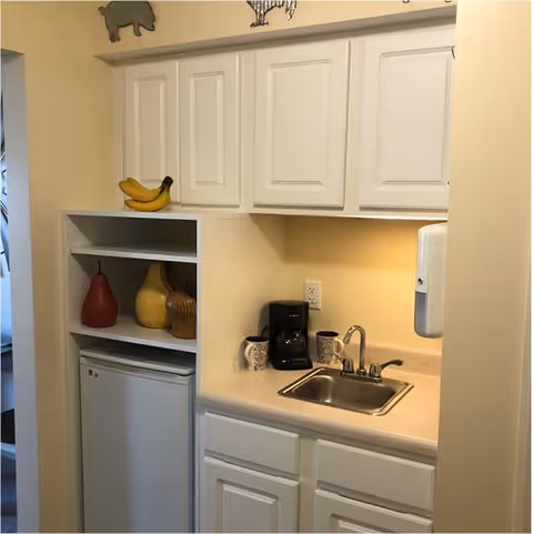 Small kitchenette area with white cabinets, a countertop with a sink, a coffee maker, two mugs, and a soap dispenser mounted on the wall. There is a mini refrigerator below open shelves holding decorative items including a red pear, a yellow pear, and a basket. A bunch of bananas is placed on top of the upper cabinets.