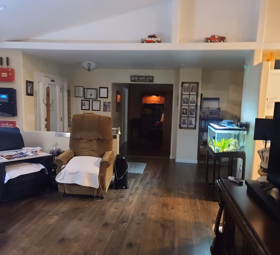 Interior view of a living room area in an assisted living facility with wooden flooring, a brown recliner chair with a white cloth draped over it, a black armchair with a white cloth, a fish tank on a stand, framed pictures and certificates on the walls, and a hallway leading to another room.