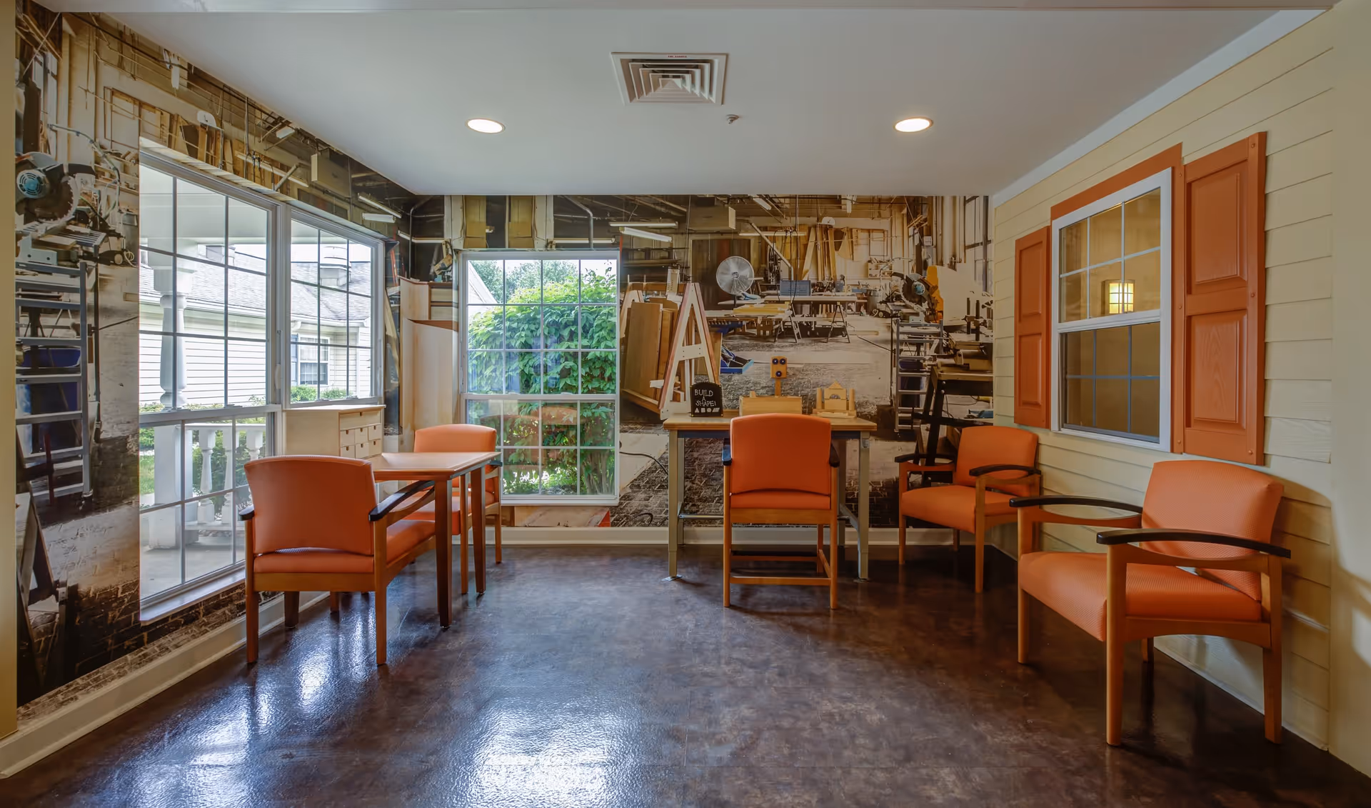 A small seating area with four orange chairs and two small tables in a room with large windows. The walls feature a mural of a workshop with tools and equipment, and one wall has a window with orange shutters. The floor is dark and shiny, and the ceiling has recessed lighting.