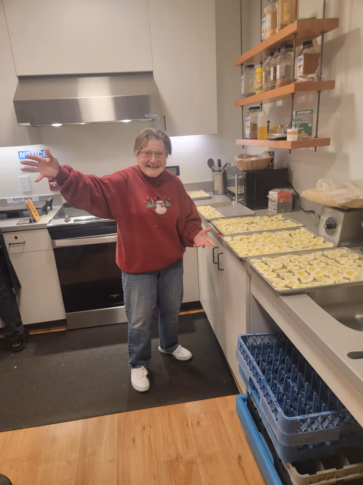 An elderly woman wearing a red sweater with a snowman design and jeans stands in a kitchen smiling with her arms open. Several trays filled with halved boiled eggs are placed on the kitchen counter beside her. The kitchen has white cabinets, a stainless steel stove, and shelves with various jars and containers.