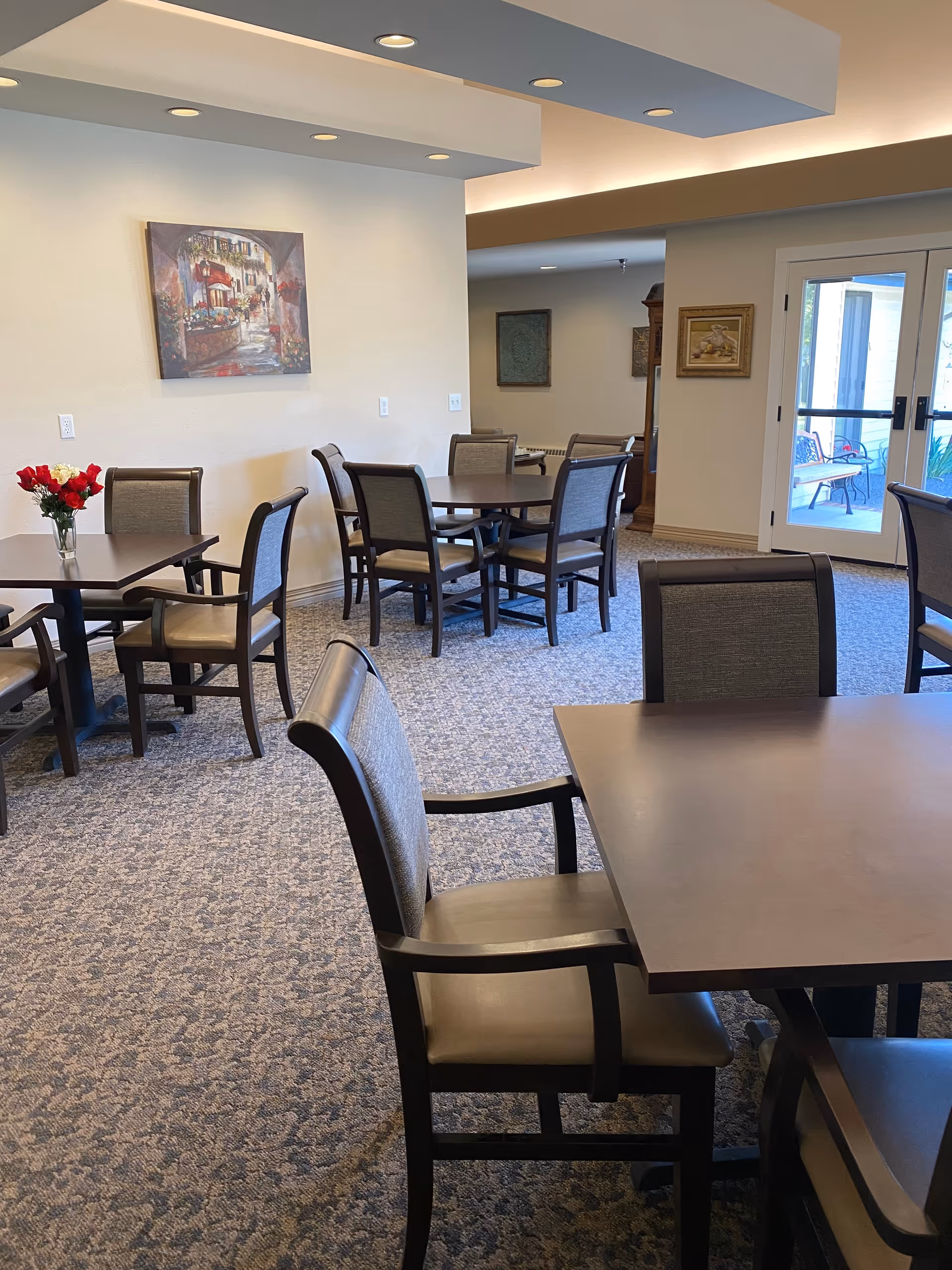 Interior view of a dining area in a retirement community with several tables and chairs arranged on a carpeted floor. A vase with red flowers is placed on one table. The walls are light-colored with framed artwork, and there are glass doors leading to an outdoor patio area with seating.