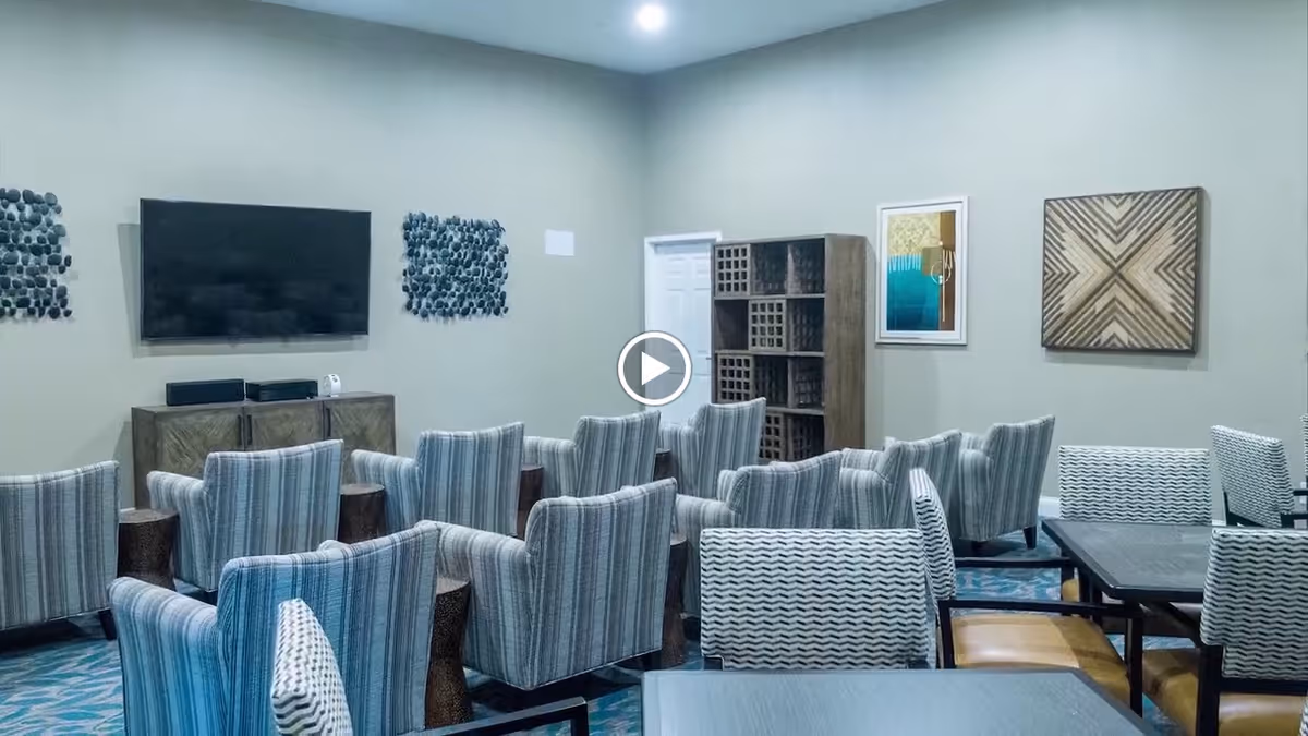 A common area with multiple upholstered armchairs arranged in rows facing a wall-mounted flat screen TV. The room has neutral-colored walls decorated with abstract art pieces and a wooden shelving unit. There are also tables with chairs in the foreground.