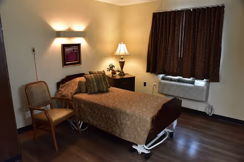 A small, neatly arranged bedroom in an assisted living facility featuring a single bed with a brown patterned bedspread and two pillows. Next to the bed is a wooden nightstand with a lamp and a small plant. A wooden chair with a cushioned seat is placed beside the bed. The room has dark wood flooring, a window with closed brown curtains, and a wall-mounted air conditioning unit below the window. A framed picture hangs on the wall above the bed, illuminated by a wall light.
