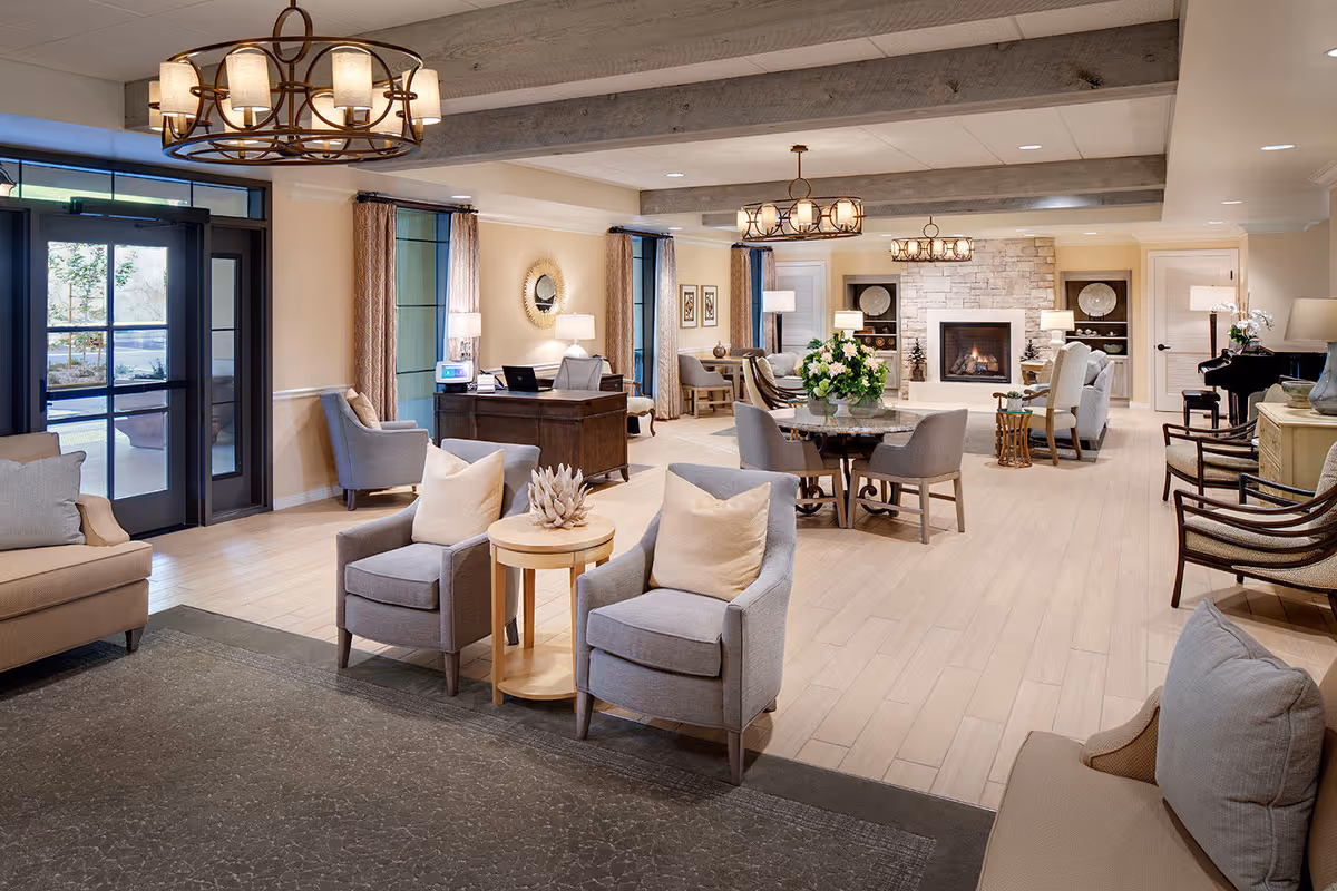 A spacious and elegantly furnished senior living common area featuring multiple seating arrangements with armchairs and sofas, a round table with chairs, a stone fireplace, decorative lighting fixtures, large windows with curtains, and a piano in the corner.