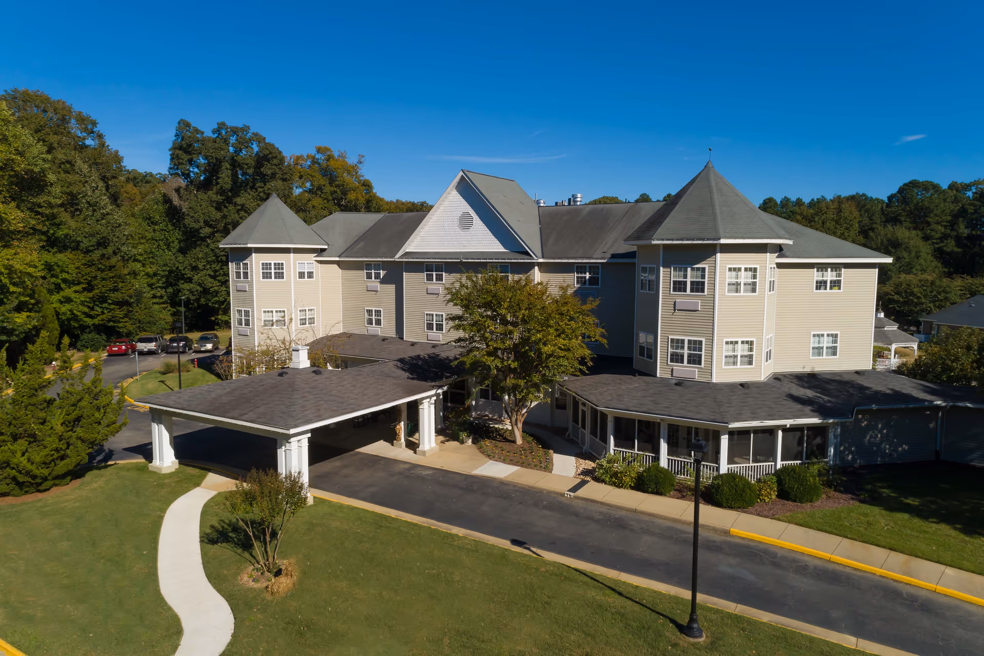 Aerial view of Mennowood Retirement Community building with a covered entrance, surrounded by green lawns, trees, and a parking area under a clear blue sky.