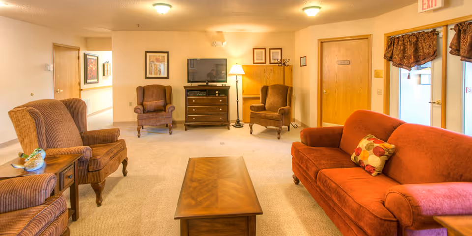 A warm common living room with upholstered armchairs, a red sofa, wooden coffee table, TV and lamp in a senior living facility.