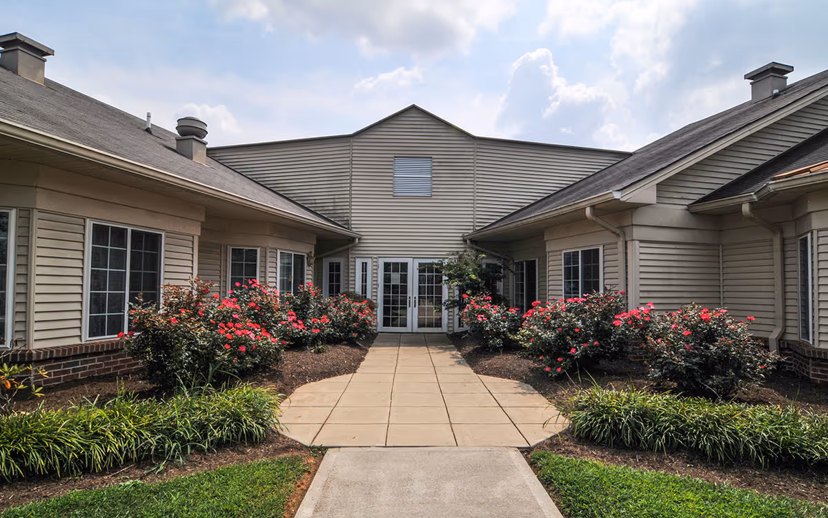 Exterior view of a senior living facility building with beige siding, multiple windows, and a central double glass door entrance. The entrance pathway is flanked by well-maintained flower beds with blooming red flowers and green shrubs under a partly cloudy sky.