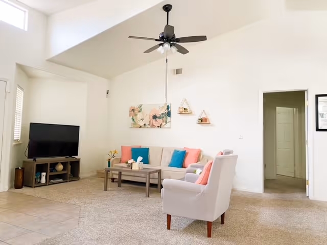 A bright living room with a beige sofa adorned with pink and blue cushions, a matching armchair with a pink cushion, a wooden coffee table, and a TV on a stand. The room has a ceiling fan, light-colored walls, carpeted floor, and decorative wall art above the sofa.