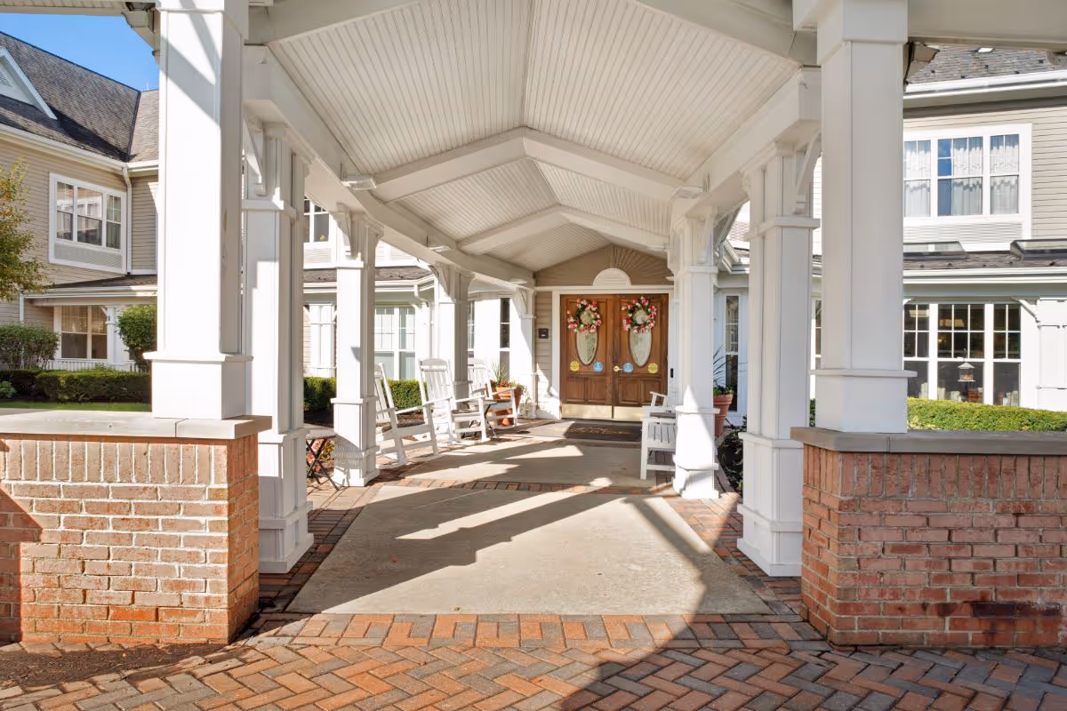 Covered entrance walkway to a senior living facility with white columns, brick half-walls, and several white rocking chairs along the sides. Double wooden doors decorated with floral wreaths are visible at the end of the walkway. The building exterior is light-colored with multiple windows.