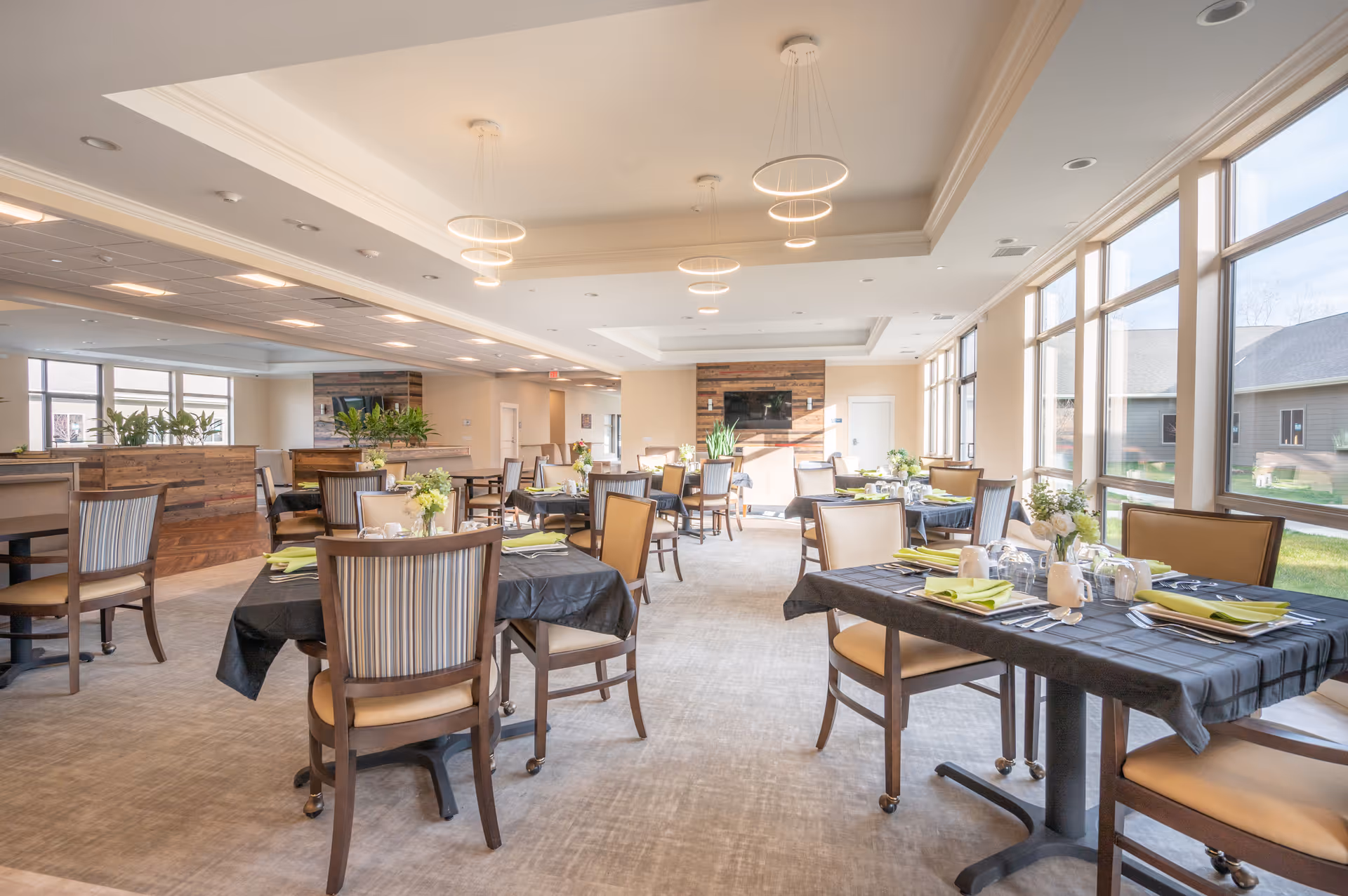 Bright and spacious dining room with multiple tables covered in black tablecloths, each set with green napkins, plates, and glassware. The room features large windows letting in natural light, modern circular ceiling lights, and a cozy seating area with a TV mounted on a wooden accent wall in the background.