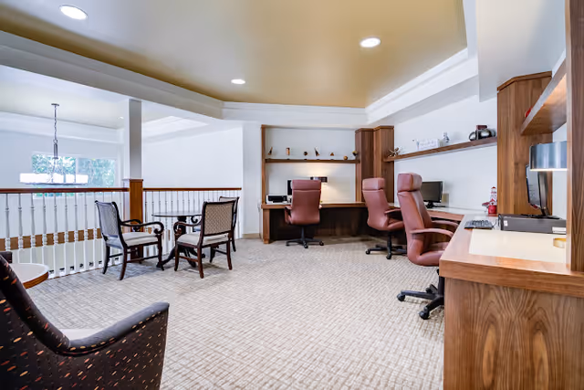 A spacious interior room with a beige carpet and white walls, featuring a row of desks with computers and brown office chairs along one wall. There are additional chairs and a small table near a railing overlooking a lower level, with recessed ceiling lights providing illumination.