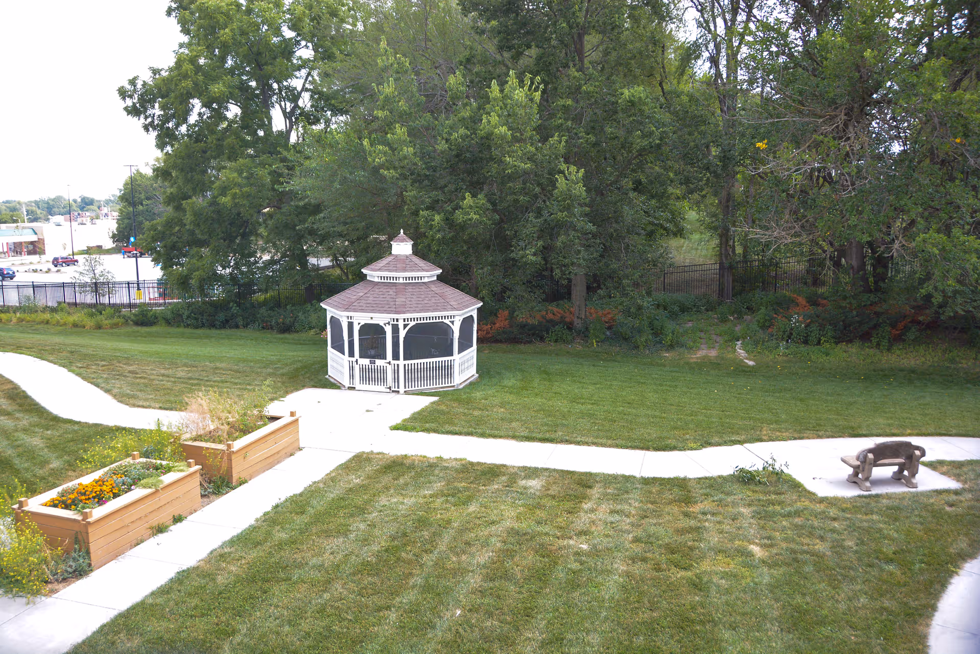 A well-maintained outdoor garden area with a white gazebo, green grass, a wooden bench on a concrete pad, and two raised garden beds with flowers and plants. Trees and shrubs form a natural backdrop.