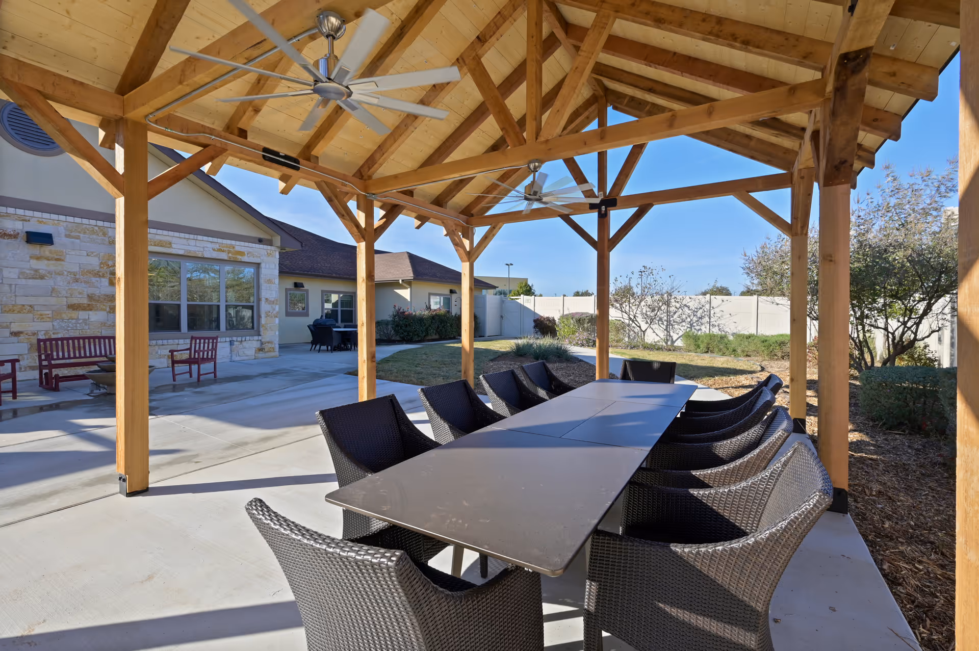 Covered outdoor pavilion with a long dining table and wicker chairs beneath a wooden vaulted roof.