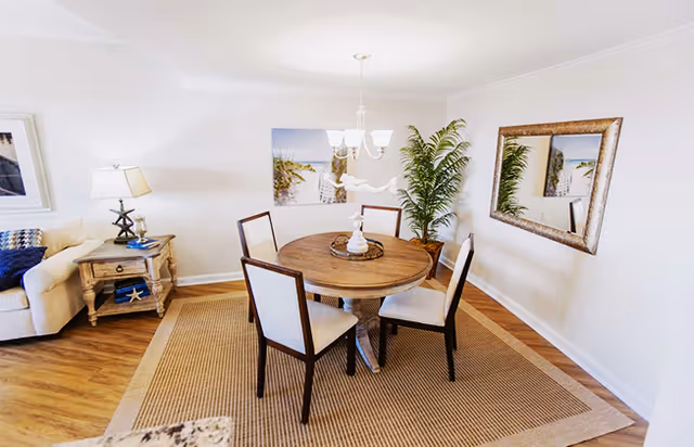 A bright dining area featuring a round wooden table with four cushioned chairs on a large woven rug. The room has light-colored walls with a large mirror and a framed beach-themed picture. A potted plant is placed in the corner, and a beige sofa with a side table and lamp is partially visible on the left side.