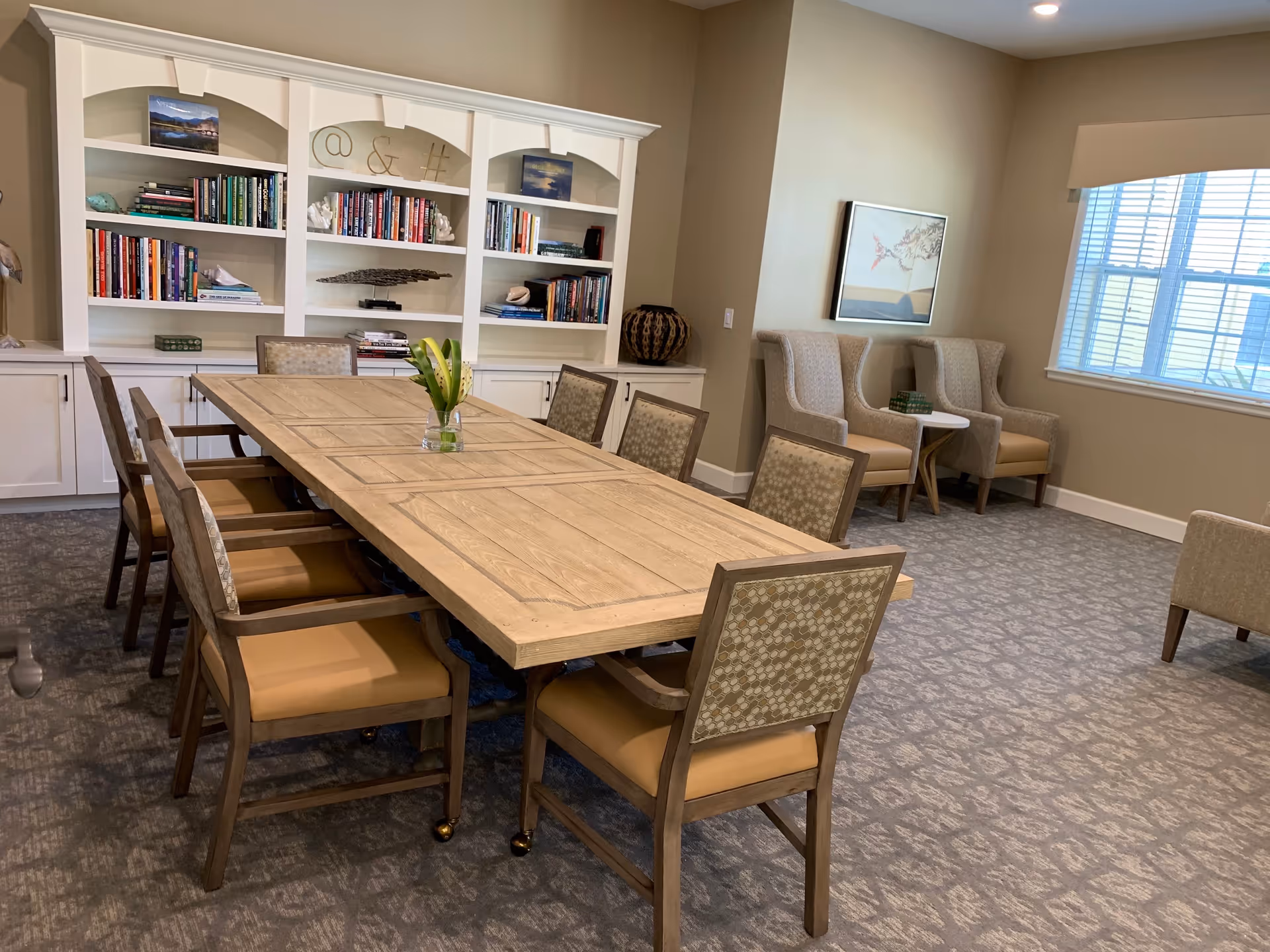 A well-lit interior room with a long wooden table surrounded by eight cushioned chairs. Behind the table is a white built-in bookshelf filled with books and decorative items. To the right, there are three upholstered armchairs arranged near a window with blinds, and a small side table with books. The room has beige walls and patterned carpet flooring.