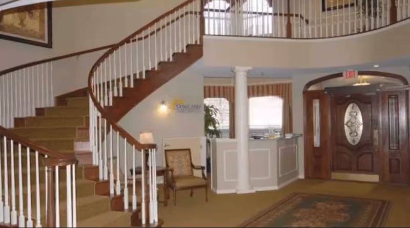 Interior lobby with a curved staircase, reception desk, front door, chairs, and a decorative rug.