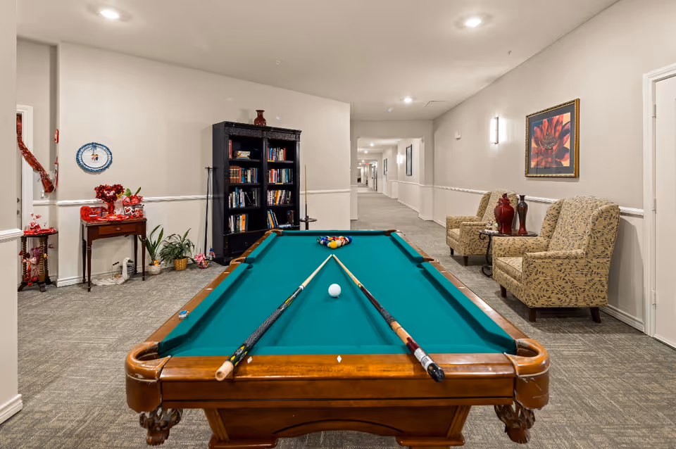 Indoor recreational area with a pool table in the center, two pool cues resting on the table, a bookshelf filled with books, two patterned armchairs with a small table holding decorative vases, and a long hallway extending into the background. The walls are light-colored with framed artwork and wall sconces providing lighting.