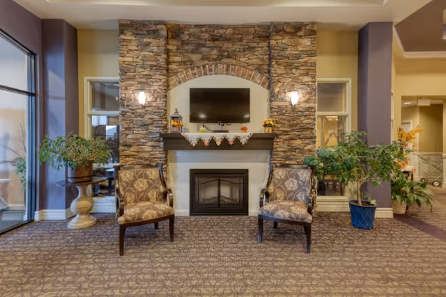 A cozy senior living facility common area featuring a stone fireplace with a mounted flat-screen TV above it. Two patterned armchairs are placed symmetrically on either side of the fireplace. There are potted plants on both sides, and the room has warm lighting with wall sconces flanking the fireplace. Large windows and an open hallway are visible in the background.