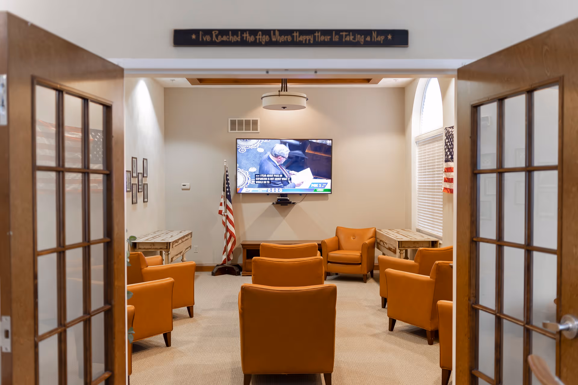 View through open double wooden doors into a cozy common room with six orange armchairs arranged facing a wall-mounted television. An American flag stands in the corner, and two small rustic tables are placed against the side walls. Above the doorway is a sign that reads, 'I've Reached the Age Where Happy Hour Is Taking a Nap.'