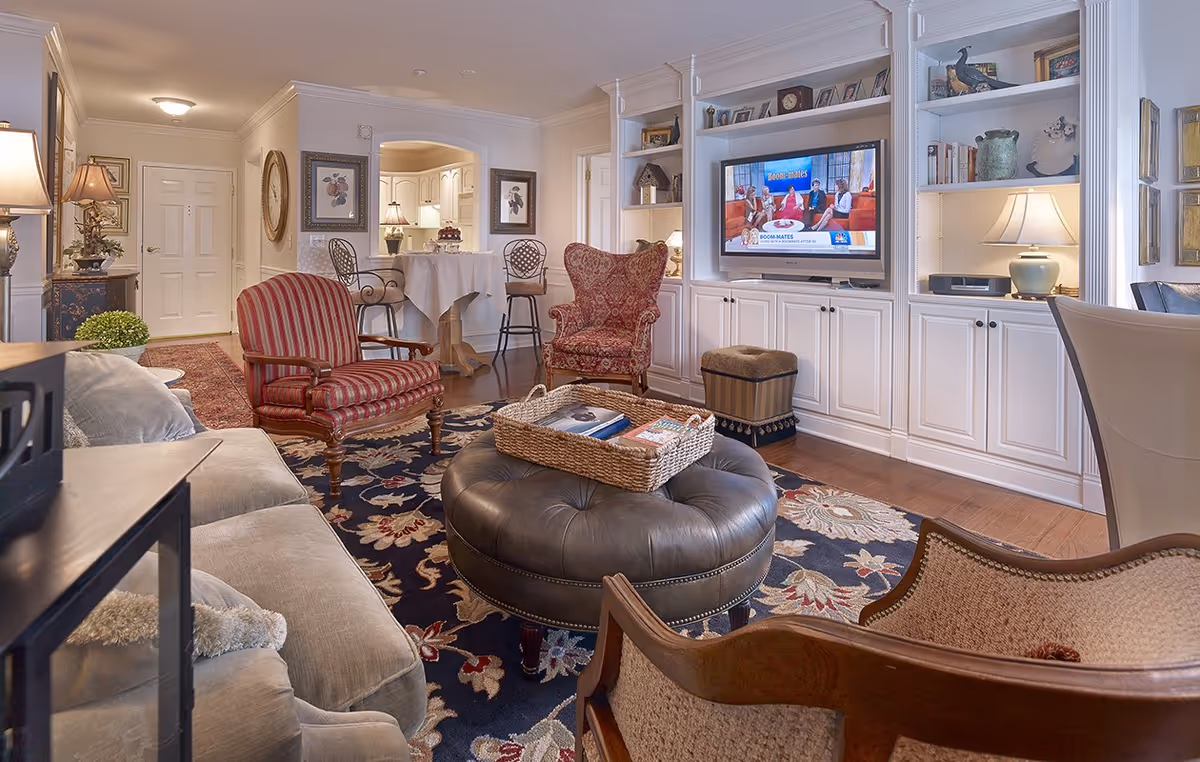 A cozy living room with a beige sofa, a red striped armchair, a patterned wingback chair, and a round leather ottoman with a wicker tray on top. The room features a dark floral rug, built-in white shelves and cabinets with a TV displaying a talk show, and soft lighting from table lamps. In the background, there is a small dining area with two bar stools and a table covered with a white tablecloth.
