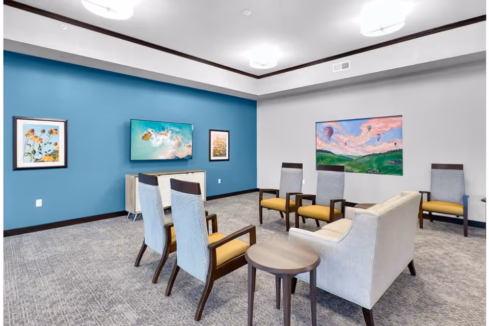 Bright communal lounge with chairs and a sofa arranged facing a wall-mounted TV and colorful artwork on the walls.