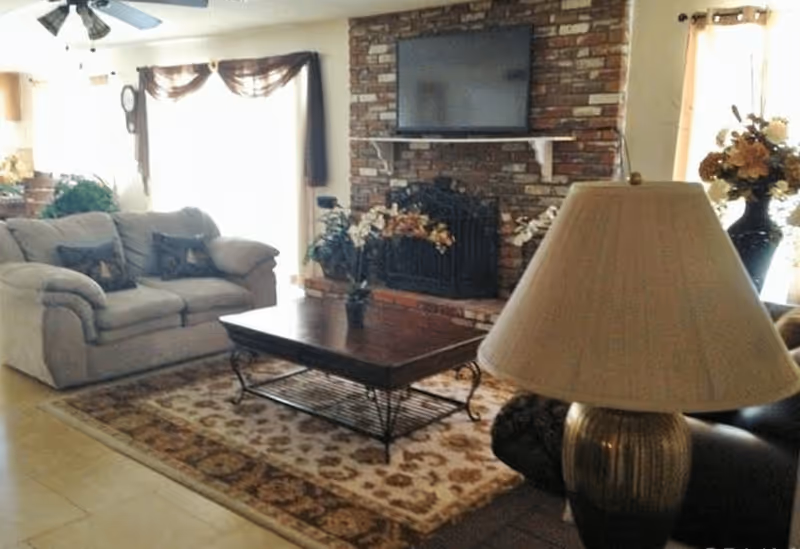 A cozy living room with a beige couch, a wooden coffee table on a patterned rug, a brick fireplace with a TV mounted above it, and a table lamp in the foreground. There are floral arrangements on either side of the fireplace and large windows with curtains letting in natural light.