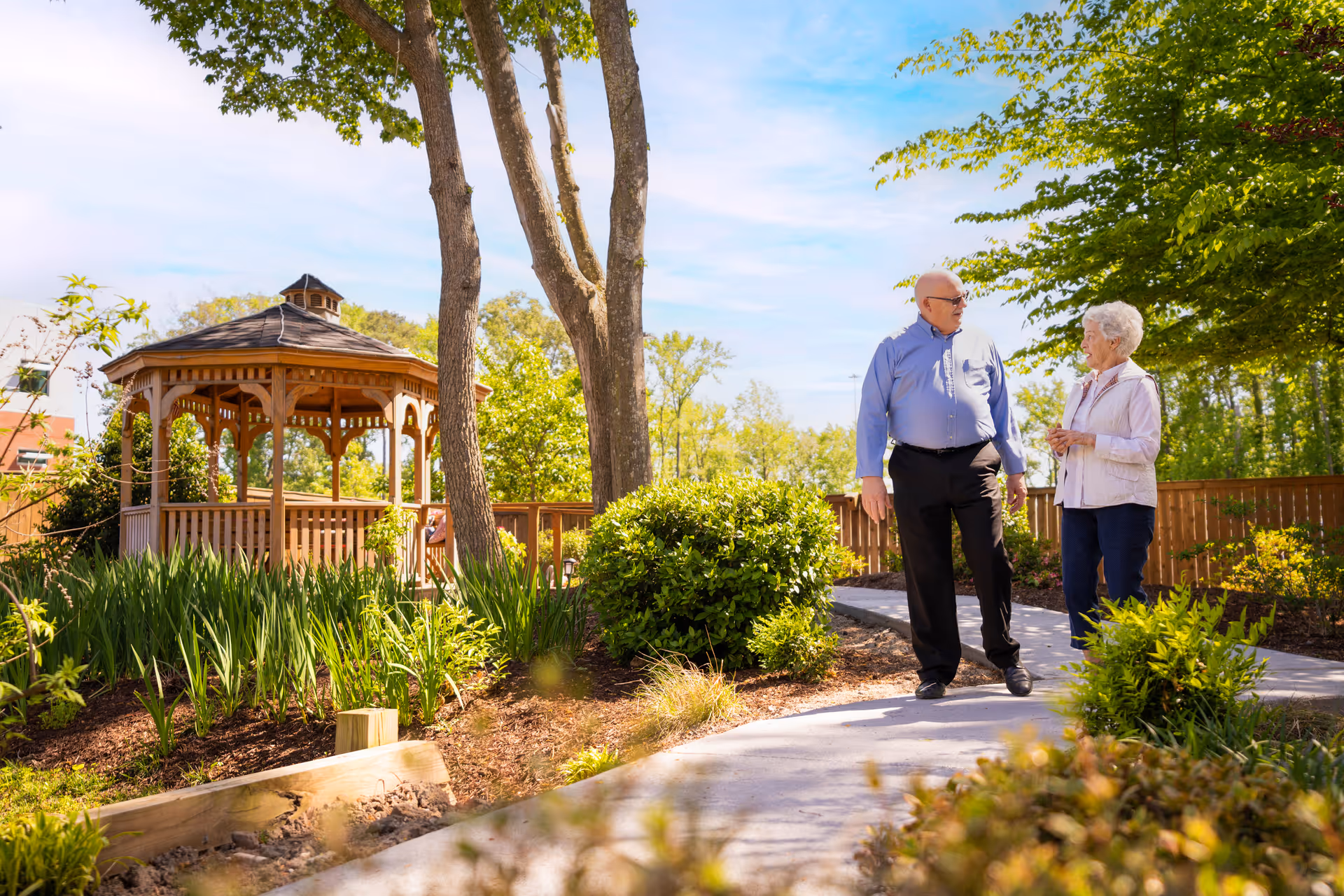 An elderly man and woman walking and talking on a paved path in a garden area with green shrubs, trees, and a wooden gazebo under a clear blue sky.