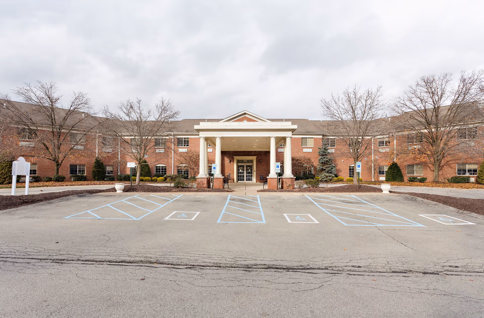 Front exterior view of a two-story brick building with a covered entrance supported by four white columns. The building is surrounded by leafless trees and landscaped bushes. There are several handicap parking spaces marked in blue in the foreground under a cloudy sky.
