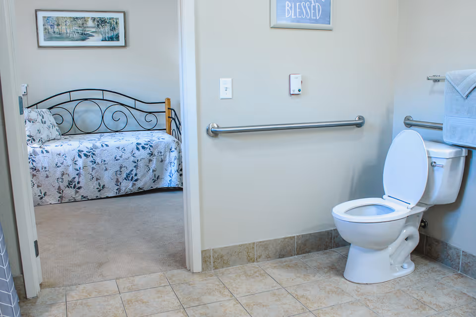 View of a bathroom with a white toilet and grab bars on the walls. A towel is hanging on a rack above the toilet. Through an open door, a bedroom is visible with a daybed covered in a floral quilt and a framed landscape painting on the wall.