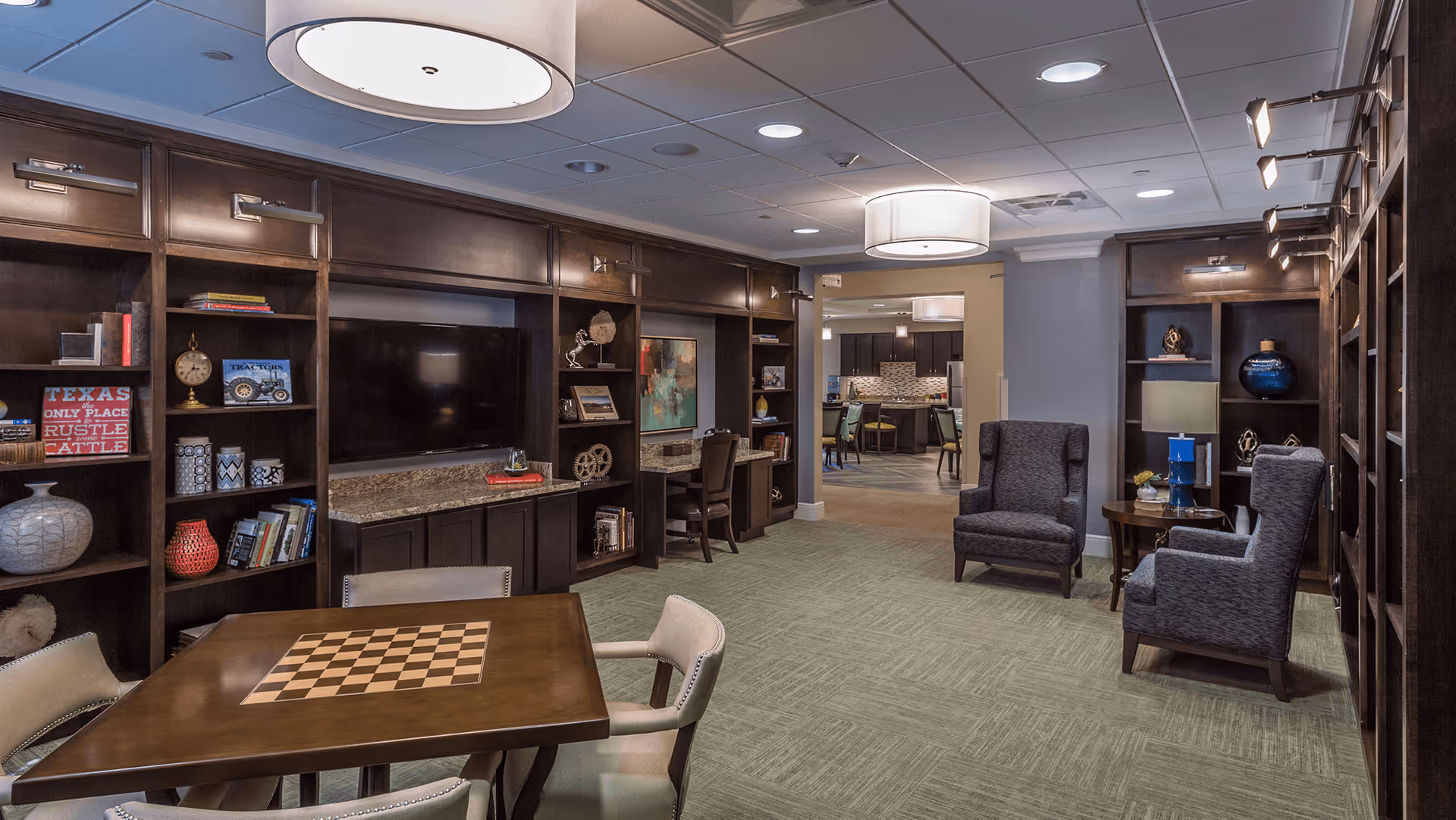 A cozy common area in a senior living facility featuring dark wood built-in bookshelves with decorative items and a large flat-screen TV. There is a wooden table with a chessboard pattern on top surrounded by chairs, and two upholstered armchairs with a small round table between them. The room has soft lighting from ceiling fixtures and leads into a kitchen or dining area in the background.