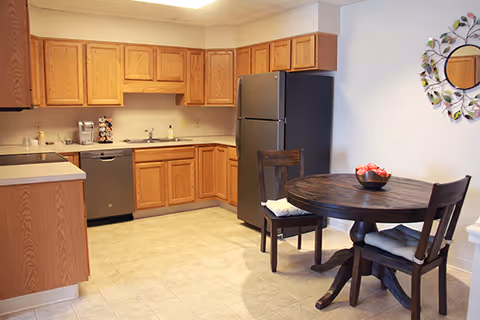 A kitchen area with wooden cabinets, a stainless steel refrigerator, dishwasher, and sink. There is a round wooden dining table with two chairs, each with a cushion. A decorative mirror with a leaf design hangs on the wall above the table.