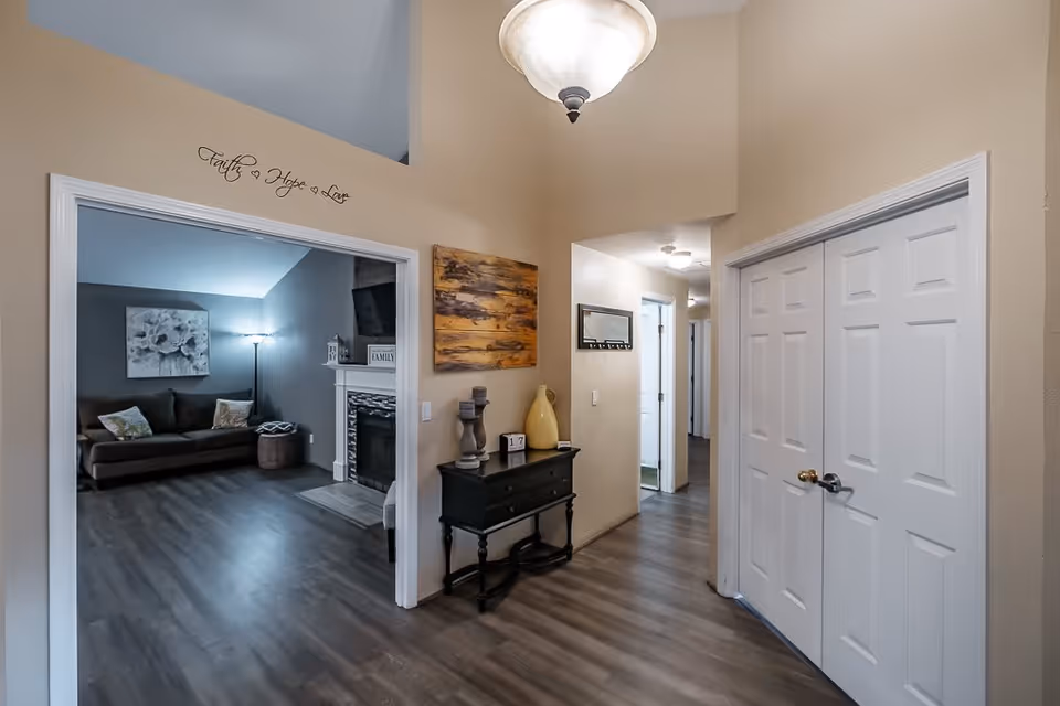 Interior view of a hallway in a residential facility named Juliet, featuring light beige walls, wood flooring, a small black console table with decorative items, and a doorway leading to a living room with a brown sofa, floor lamp, and a fireplace. Above the doorway is a wall decal that reads 'Faith Hope Love'.