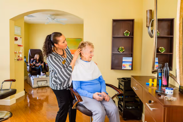 A caregiver is styling the hair of an elderly woman seated in a chair inside a well-lit room. In the background, another elderly woman is seated in a recliner chair. The room has wooden flooring, a large mirror, shelves with decorative items, and a counter with hair care products.