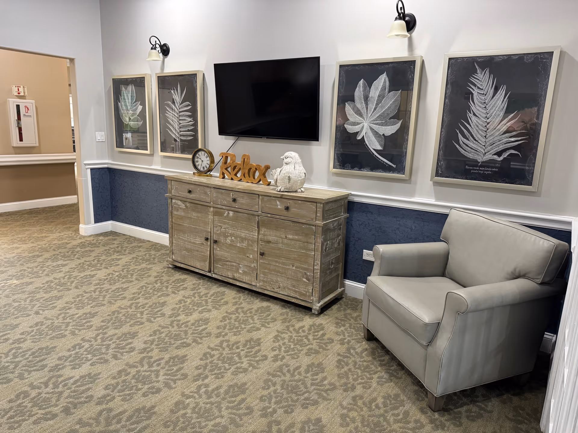 A cozy interior corner of a senior living facility featuring a gray armchair, a rustic wooden cabinet with decorative items including a clock, a 'Relax' sign, and a bird figurine. Above the cabinet is a mounted flat-screen TV and four framed botanical prints on the wall. The room has patterned carpet flooring and a two-tone wall with white upper and dark blue lower sections.