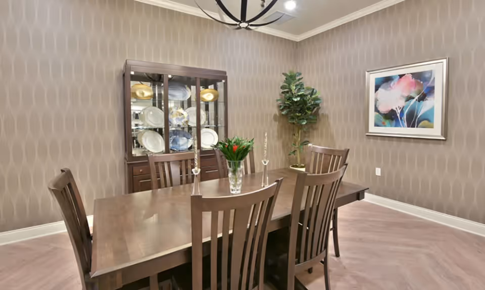 Formal dining room with a wooden table and chairs, a china cabinet, potted plant, and framed artwork on patterned walls.