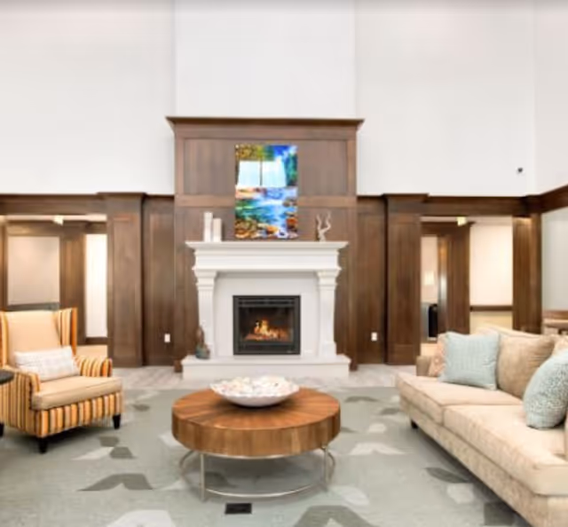 A cozy living room area with a beige sofa adorned with light blue and beige pillows, a striped armchair with a small pillow, a round wooden coffee table with a decorative bowl, and a white fireplace with a fire burning. Above the fireplace is a colorful painting. The room features dark wood paneling and light-colored walls.