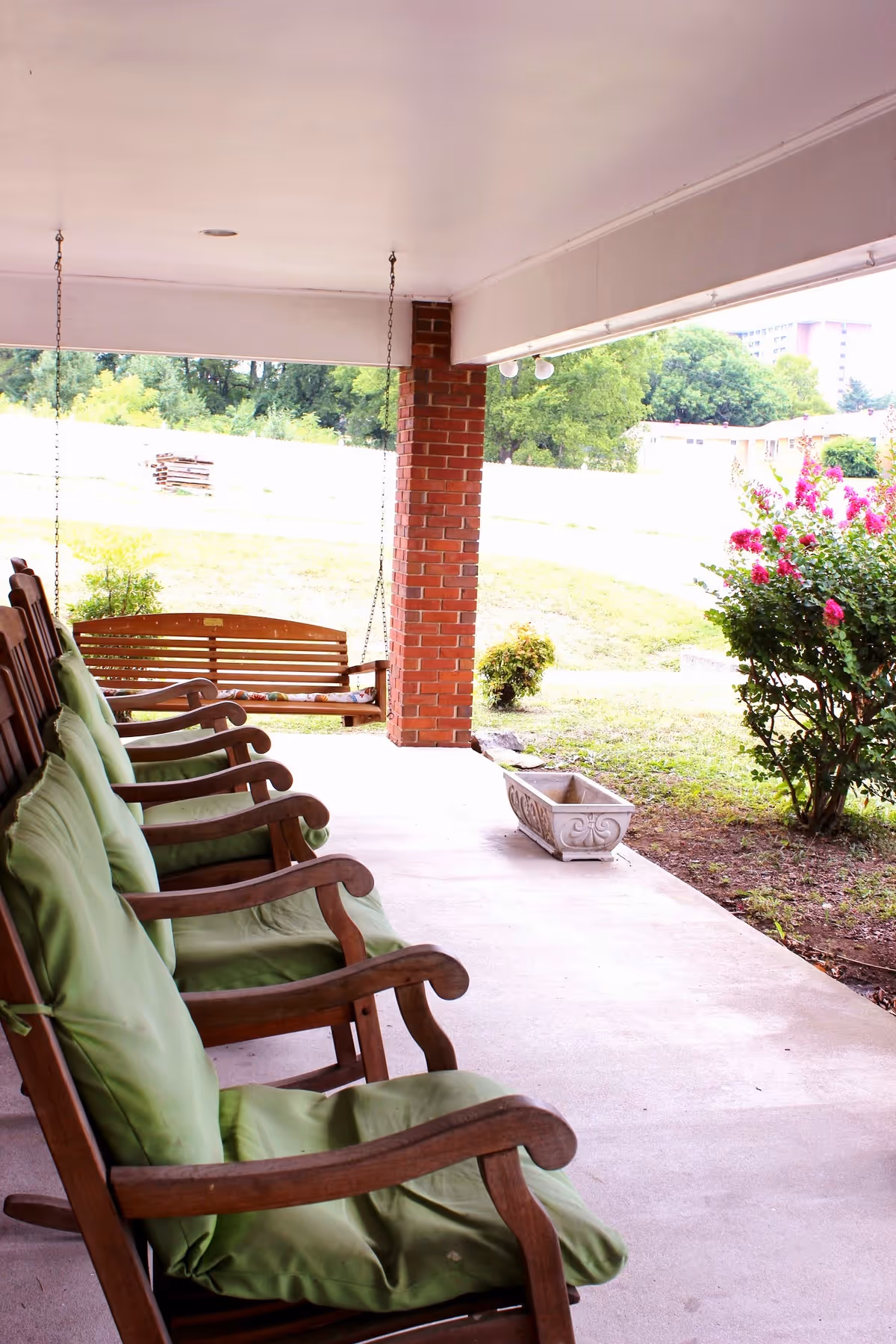 Covered outdoor patio area with wooden rocking chairs lined up, each with green cushions. A wooden porch swing hangs from the ceiling near a brick column. There is a flower planter on the ground and a flowering bush with pink flowers nearby. The background shows a grassy area with trees and buildings in the distance.