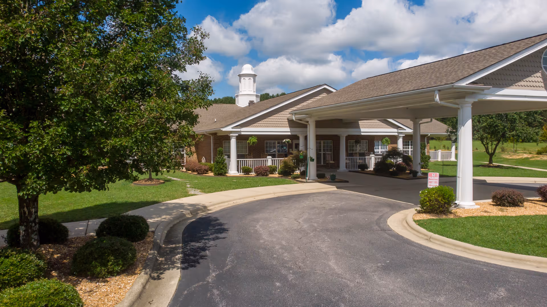 Front entrance with a covered porte-cochère, circular driveway, and landscaped lawn at a single-story brick senior living building.
