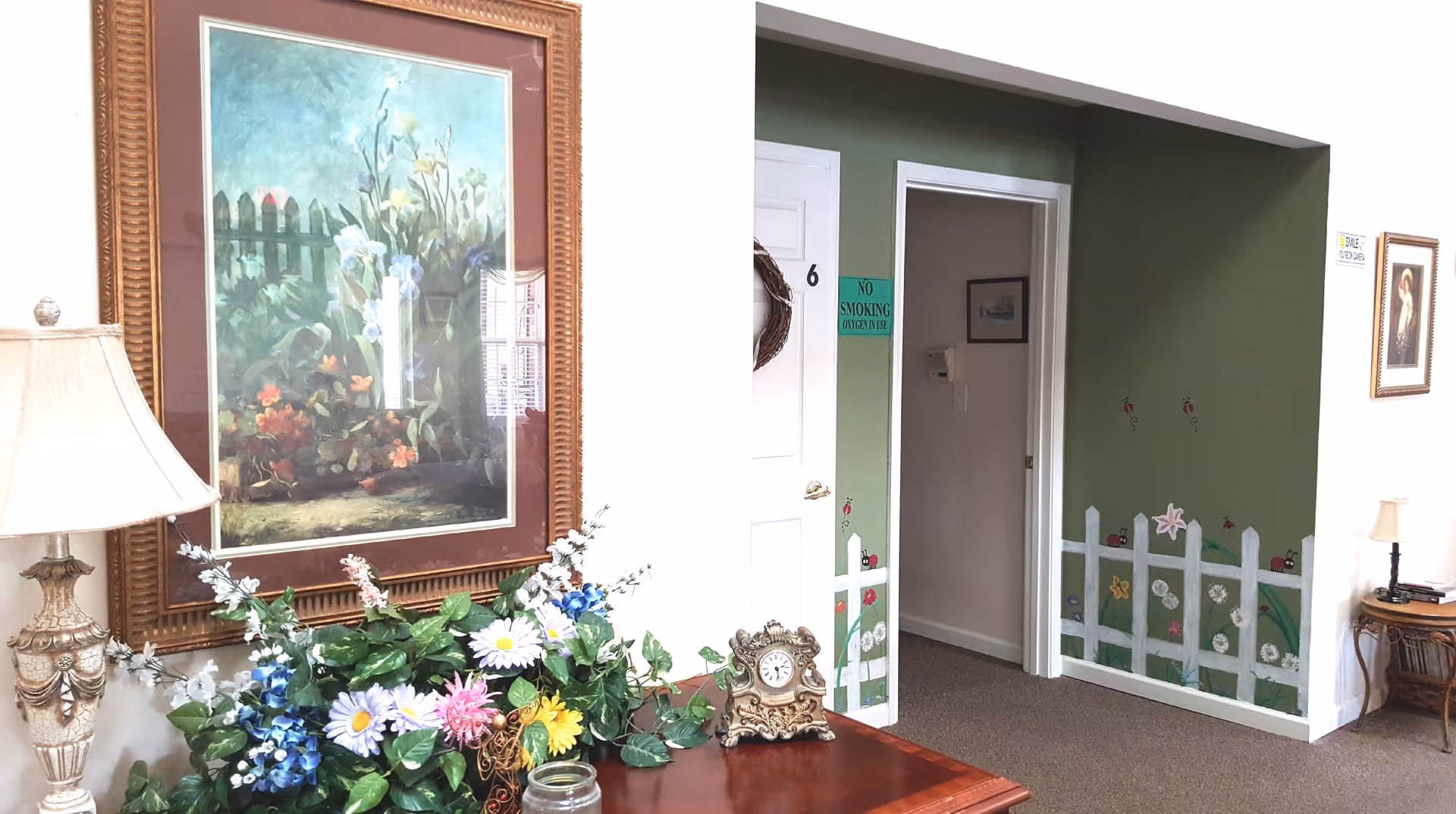 Interior view of a senior living facility room with a wooden table holding a floral arrangement, a decorative clock, and a lamp. A large framed painting of flowers and a picket fence hangs on the wall. The adjacent wall is painted green with a white picket fence mural and small ladybug and flower decorations. There is a white door marked with the number 6 and a green 'No Smoking Oxygen in Use' sign next to it. Another small table with a lamp and framed picture is visible in the background.