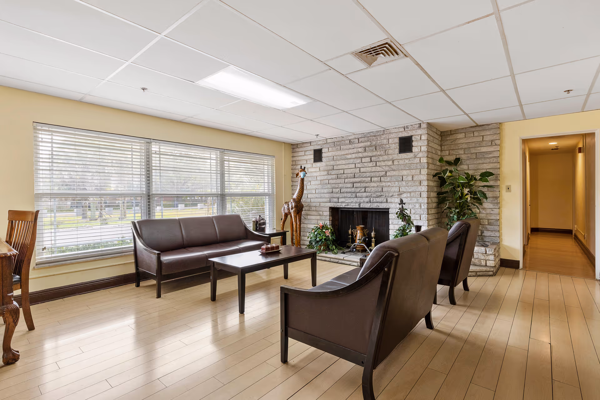 A bright living room with large windows covered by blinds, a stone fireplace, and three dark brown leather chairs arranged around a wooden coffee table. There is a tall giraffe sculpture and some potted plants near the fireplace. The floor is light wood, and there is a hallway visible to the right.