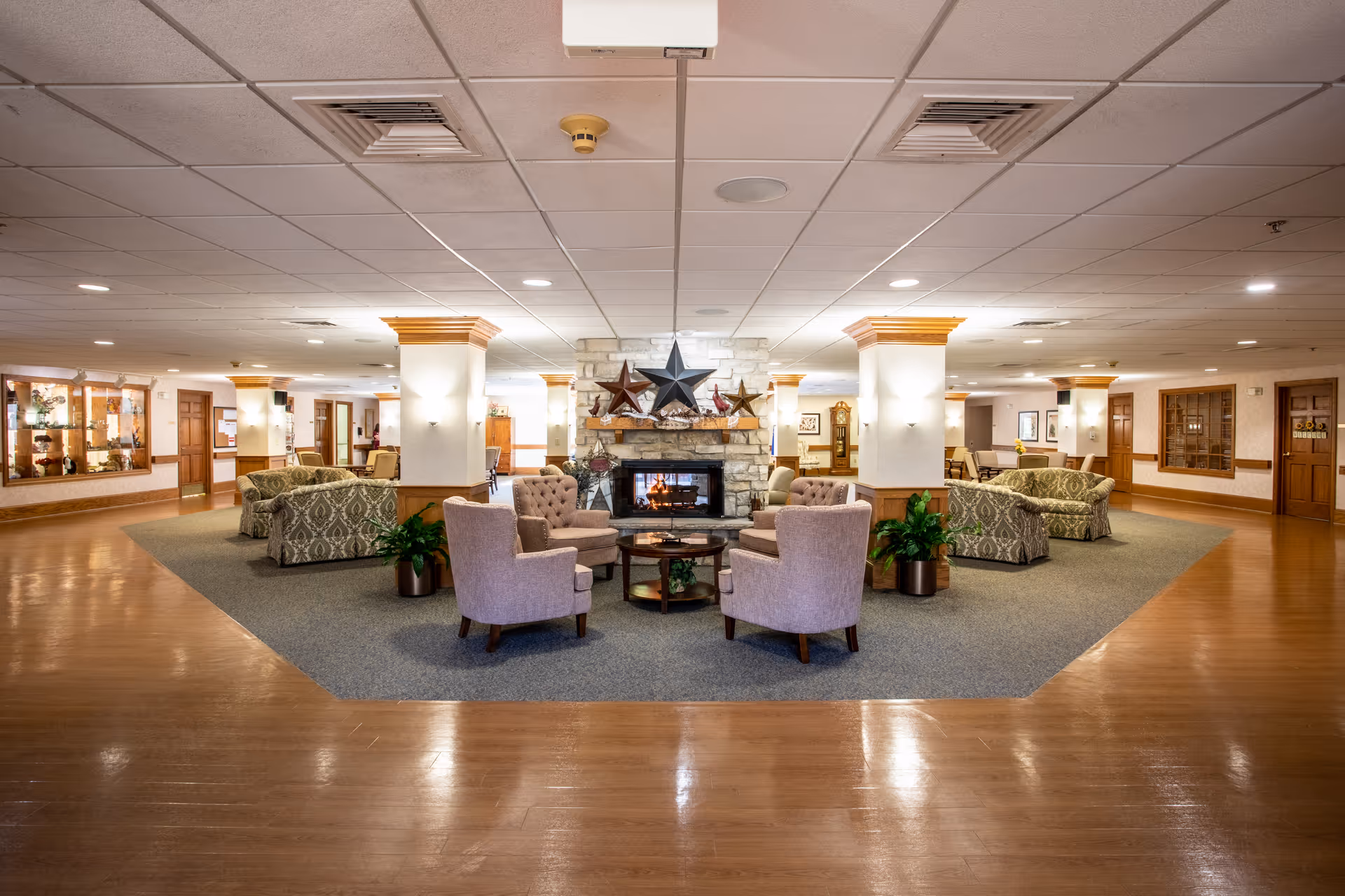 A spacious senior living community lounge area with a central stone fireplace decorated with large star ornaments. The room features multiple seating arrangements including cushioned armchairs and sofas on a carpeted area, surrounded by wooden flooring. The ceiling has recessed lighting and the walls have wooden accents and display cabinets.