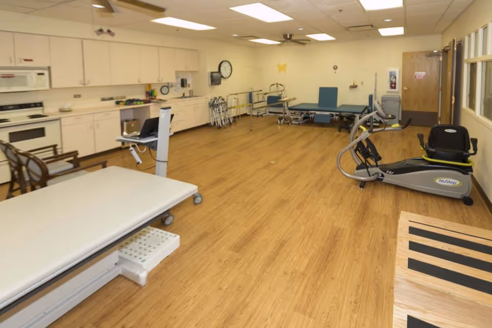 A rehabilitation therapy room with wooden flooring, equipped with exercise and therapy equipment including a NuStep recumbent cross trainer, therapy tables, walkers, chairs, and cabinets along one wall. The room has a ceiling fan, fluorescent lighting, and a clock on the wall.