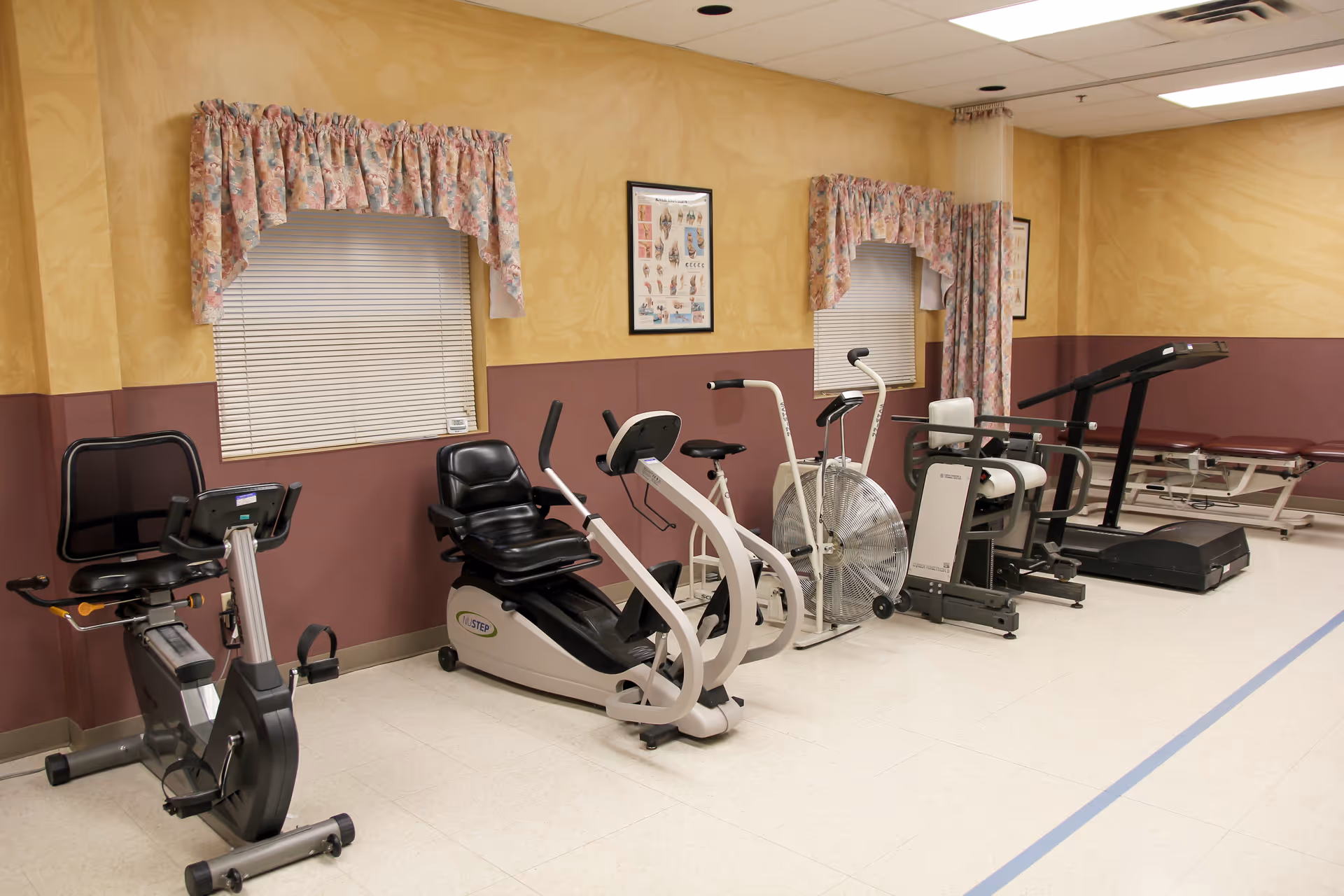A rehabilitation exercise room with various fitness equipment including stationary bikes, a fan bike, a treadmill, and a therapy table. The room has beige and maroon walls with floral curtains on the windows and framed anatomical posters on the walls.