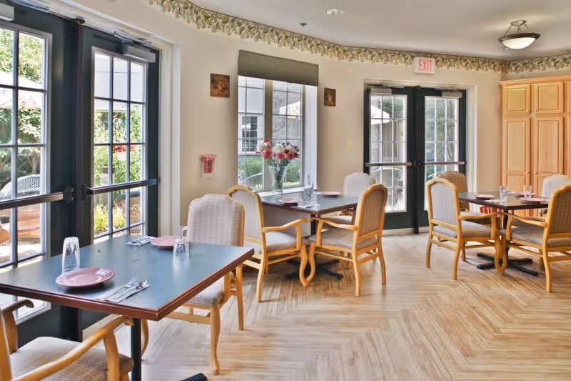 A bright dining room with several tables set with plates, glasses, and silverware. The room has large windows and glass doors letting in natural light and offering a view of an outdoor garden area. The floor has a light wood pattern, and there are cushioned wooden chairs around the tables. A vase with flowers is placed on a table near the window.