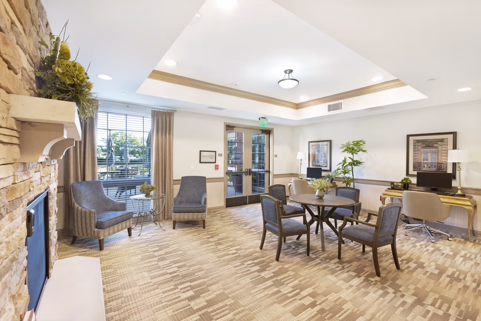 A bright and spacious common area with a stone fireplace on the left, two blue upholstered armchairs next to a window with beige curtains, a round table with four chairs in the center, and two desks with computers and beige chairs against the far wall. The room has a patterned carpet, recessed ceiling lighting, and framed artwork on the walls.